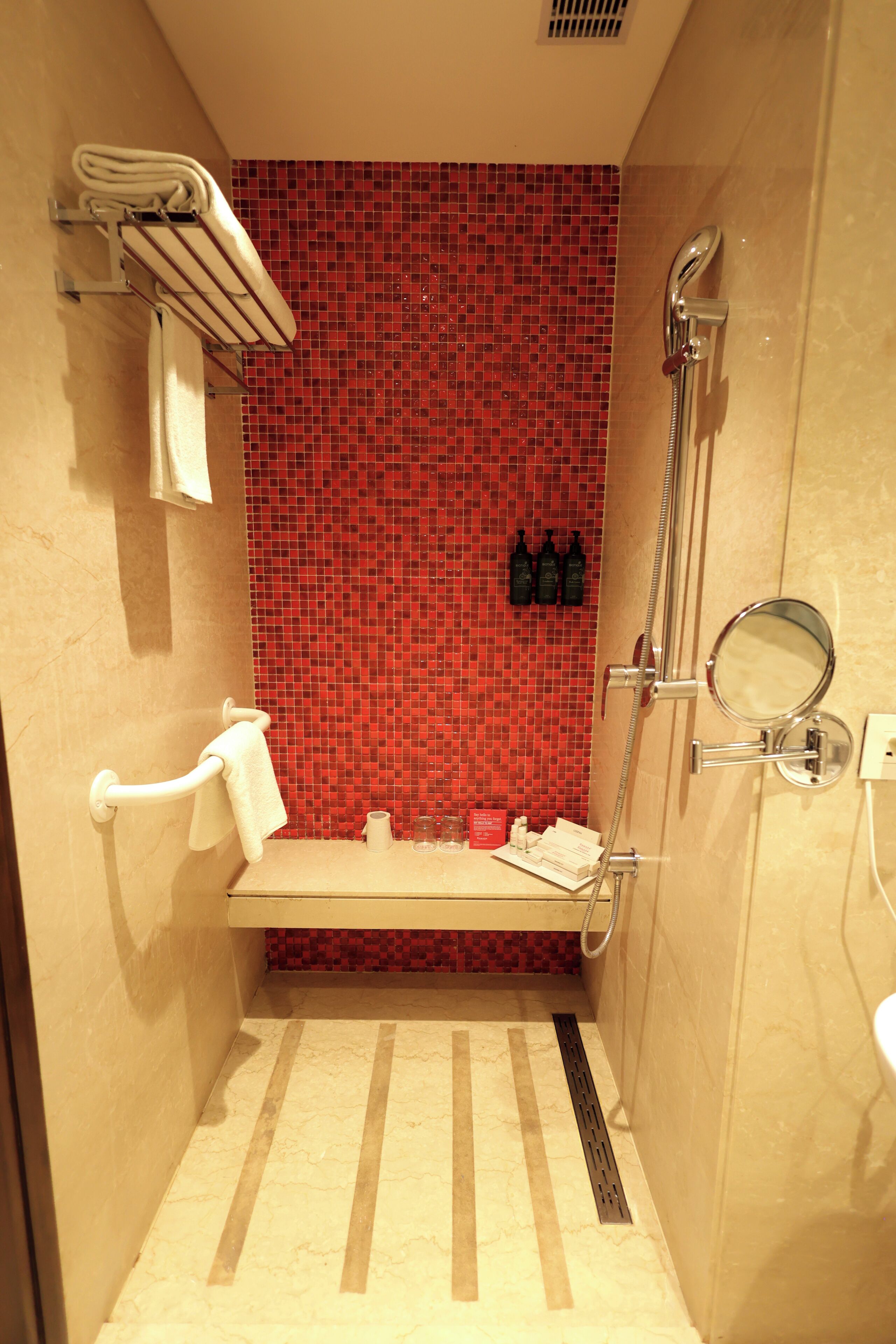 A small bathroom with a red mosaic wall, a towel rack, a mirror, and a shower.