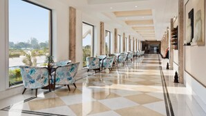 A hotel corridor with a checkered floor, large windows, and a row of chairs with floral upholstery.