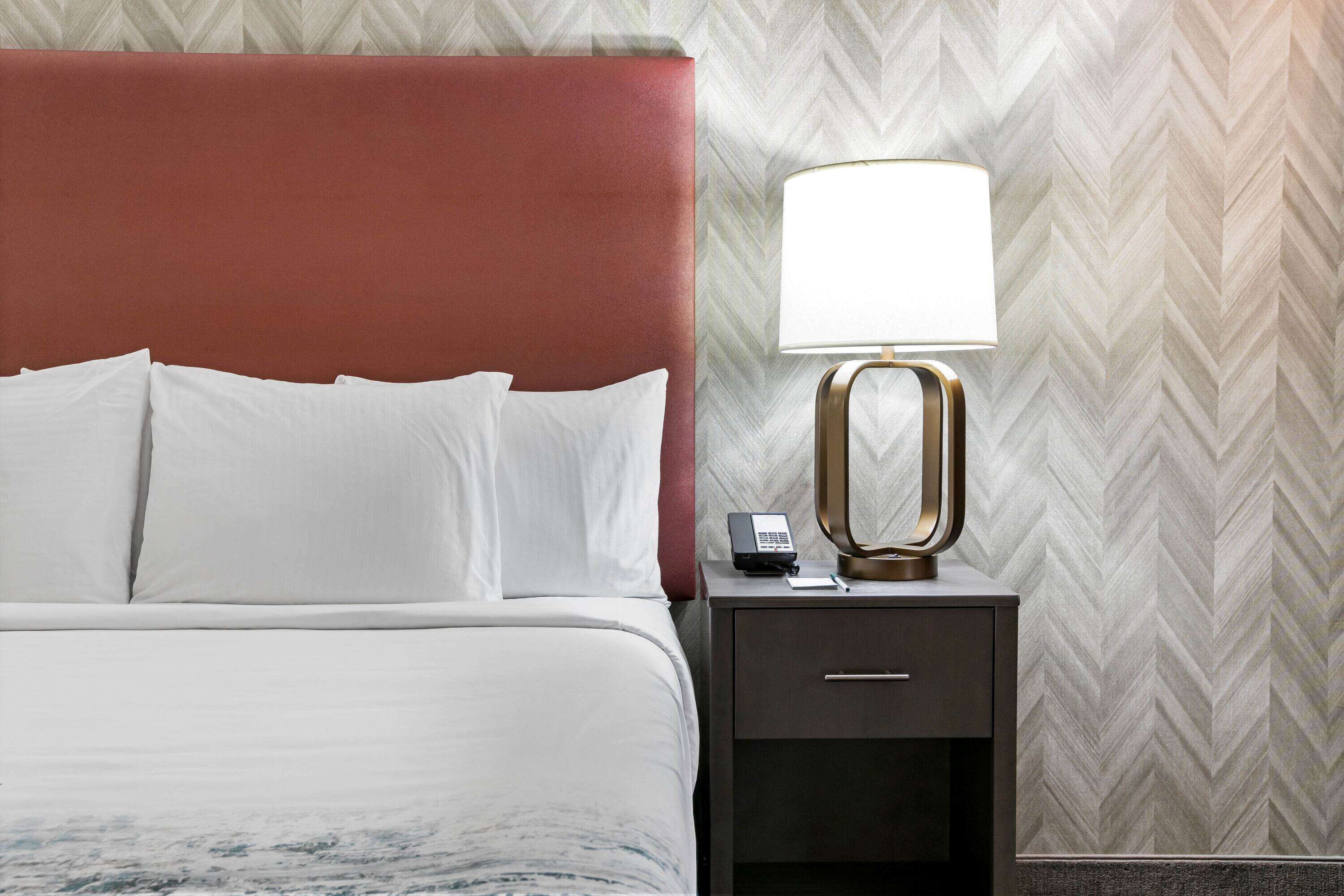 Habitación de hotel con cama, ropa de cama blanca, cabecera roja, mesita de noche con lámpara y teléfono.