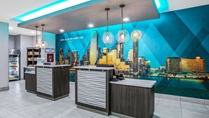 A modern hotel reception area with a cityscape mural, two reception desks, a vending machine, and a view of a body of water.