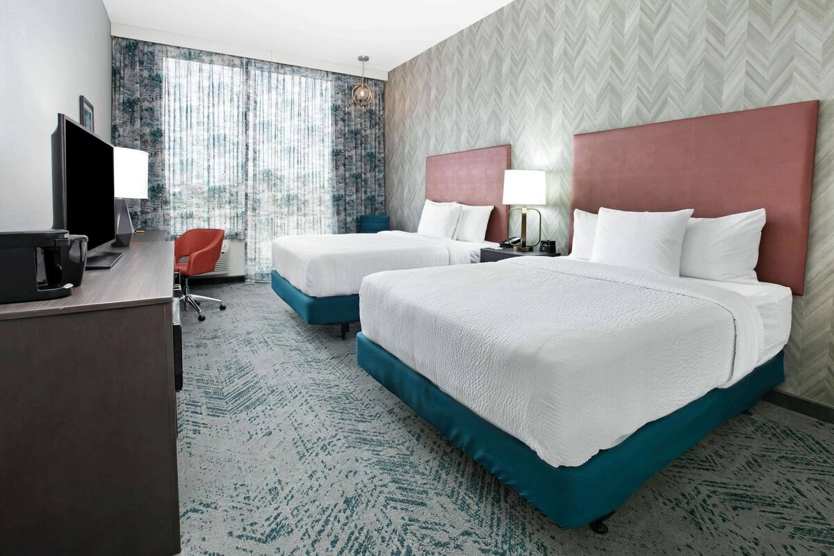 A hotel room with two beds, a desk with a computer, a chair, and a large window with curtains.