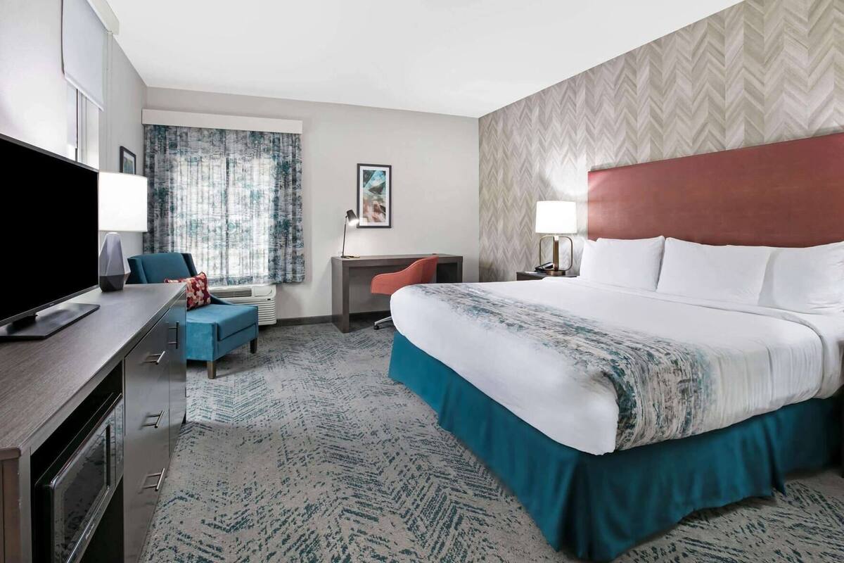 A hotel room with a bed, desk, chair, TV, and a large window with curtains.