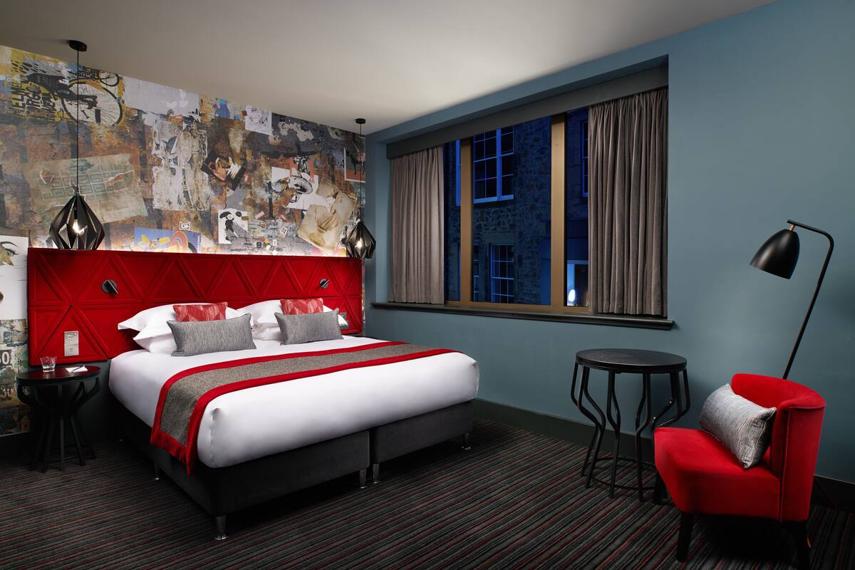 A modern hotel room with a large bed, a red accent wall, a cityscape view through the window, and a small round table.
