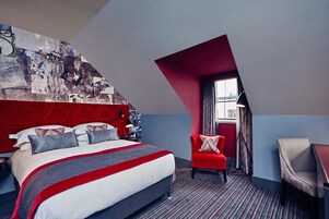 A modern hotel room with a large bed, a red accent wall, and a unique sloped ceiling design.