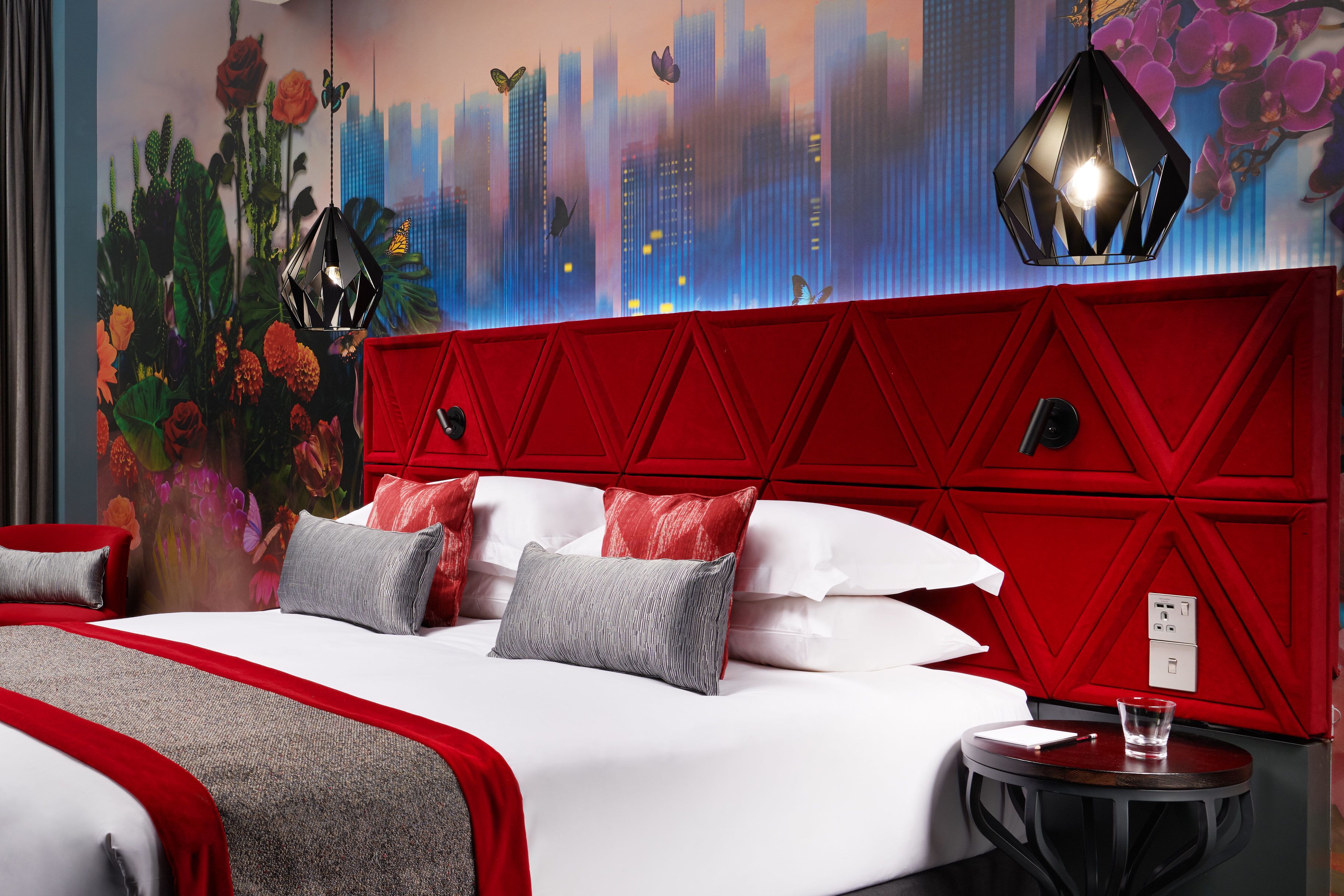 Une chambre d’hôtel avec une tête de lit rouge, un lit avec une literie blanche et grise, et une fresque représentant un paysage urbain sur le mur.