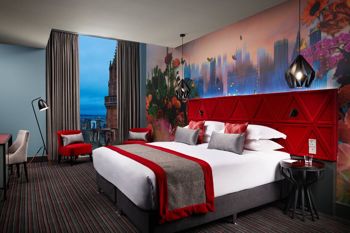 A hotel room with a large bed, a red headboard, a cityscape mural, a view of a tower, and a red accent wall.