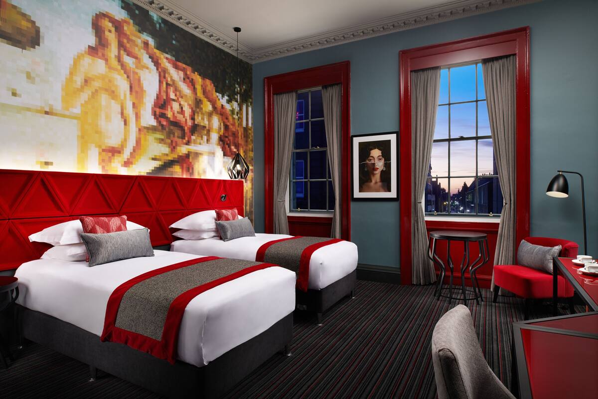 A hotel room with two beds, a red accent wall, a large painting, a city view through the window, and a desk with a chair.