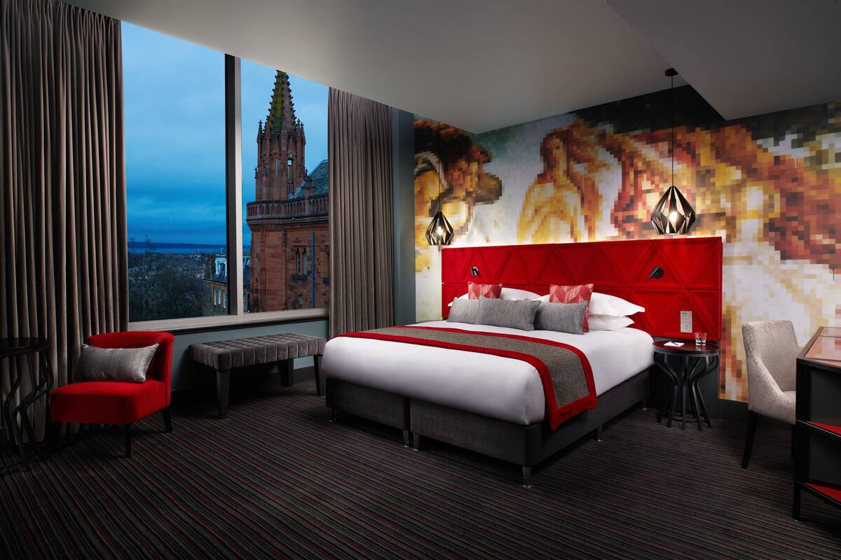 A hotel room with a large bed, a red headboard, a view of a historic building, and a modern wall mural.