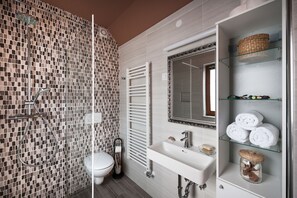 A modern bathroom with a mosaic tile wall, a glass shower enclosure, a white sink with a mirror, and shelves with towels and toiletries.