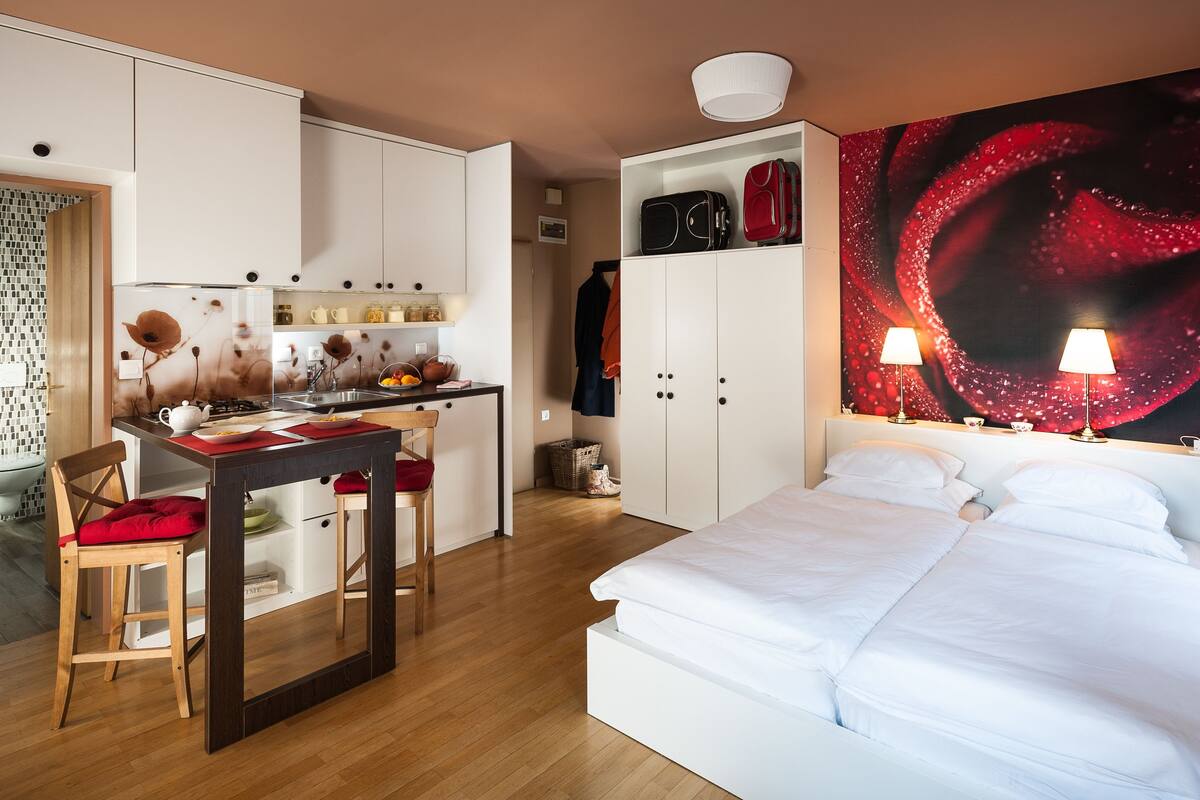 A modern hotel room with a kitchen area, a bed with white linens, and a large abstract painting on the wall.