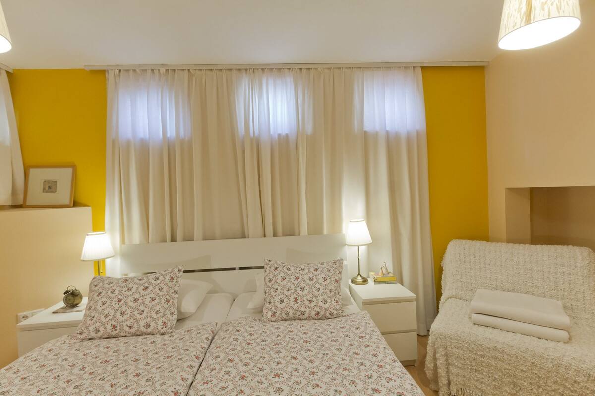 A bedroom with a bed, bedside lamps, a nightstand, a chair, and a window with curtains.