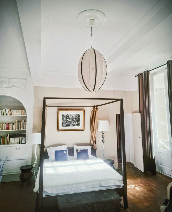 Una cama con dosel, una lámpara colgante, una estantería, un cuadro enmarcado y una ventana con cortinas.