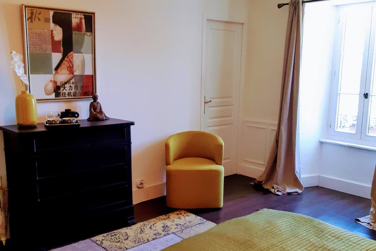 Une chambre à coucher avec un grand lit, une commode, une chaise, un lustre, une fenêtre avec des rideaux et une affiche accrochée au mur.