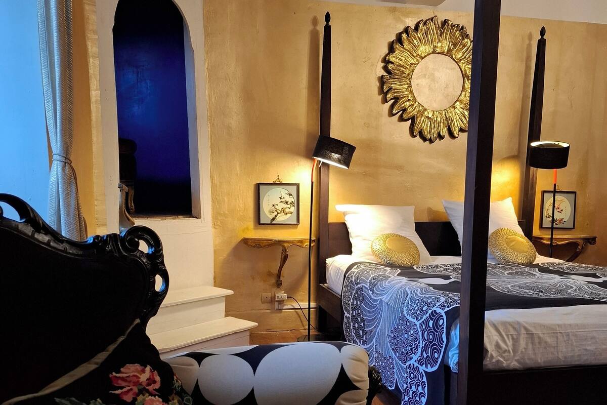 Un lit à baldaquin avec une couvre-lit à motifs bleus et blancs, une table de chevet avec une lampe et un miroir mural encadré d’or.