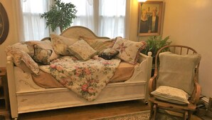 A vintage-style bedroom with a floral-patterned bedspread, a wooden chair, and a large window with sheer curtains.