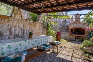 An outdoor patio with a stone wall, a wooden table with a floral tablecloth, a bench, and a brick oven.