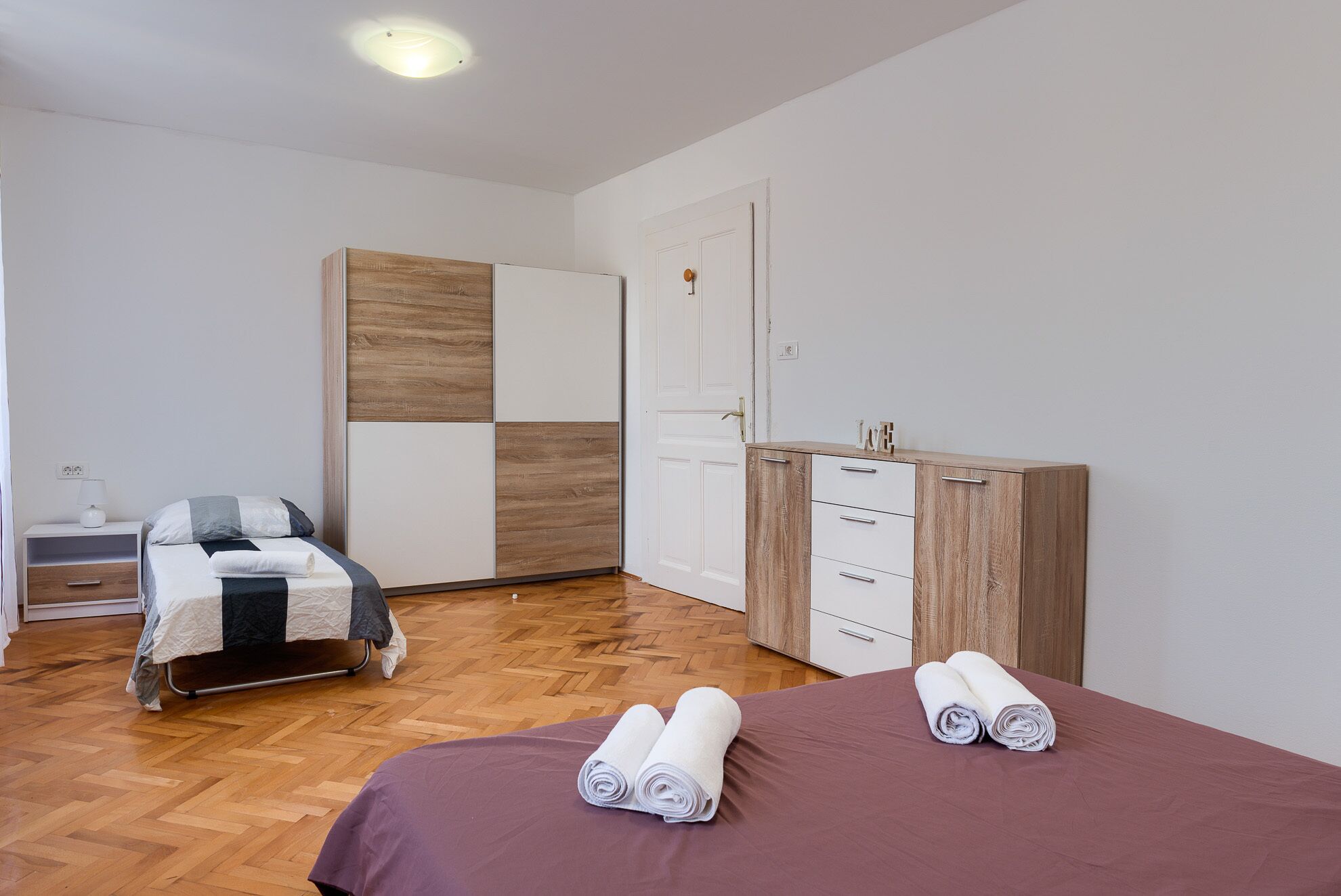 A modern bedroom with a wooden floor, a bed with rolled towels, a bedside table with a lamp, and a wardrobe with a mirror.