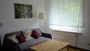 Una habitación con una mesa de madera, un sofá gris con cojines, una lámpara de pie y una ventana con cortinas translúcidas.