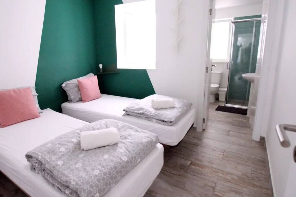 A hotel room with two beds, a green accent wall, and a bathroom visible through an open door.