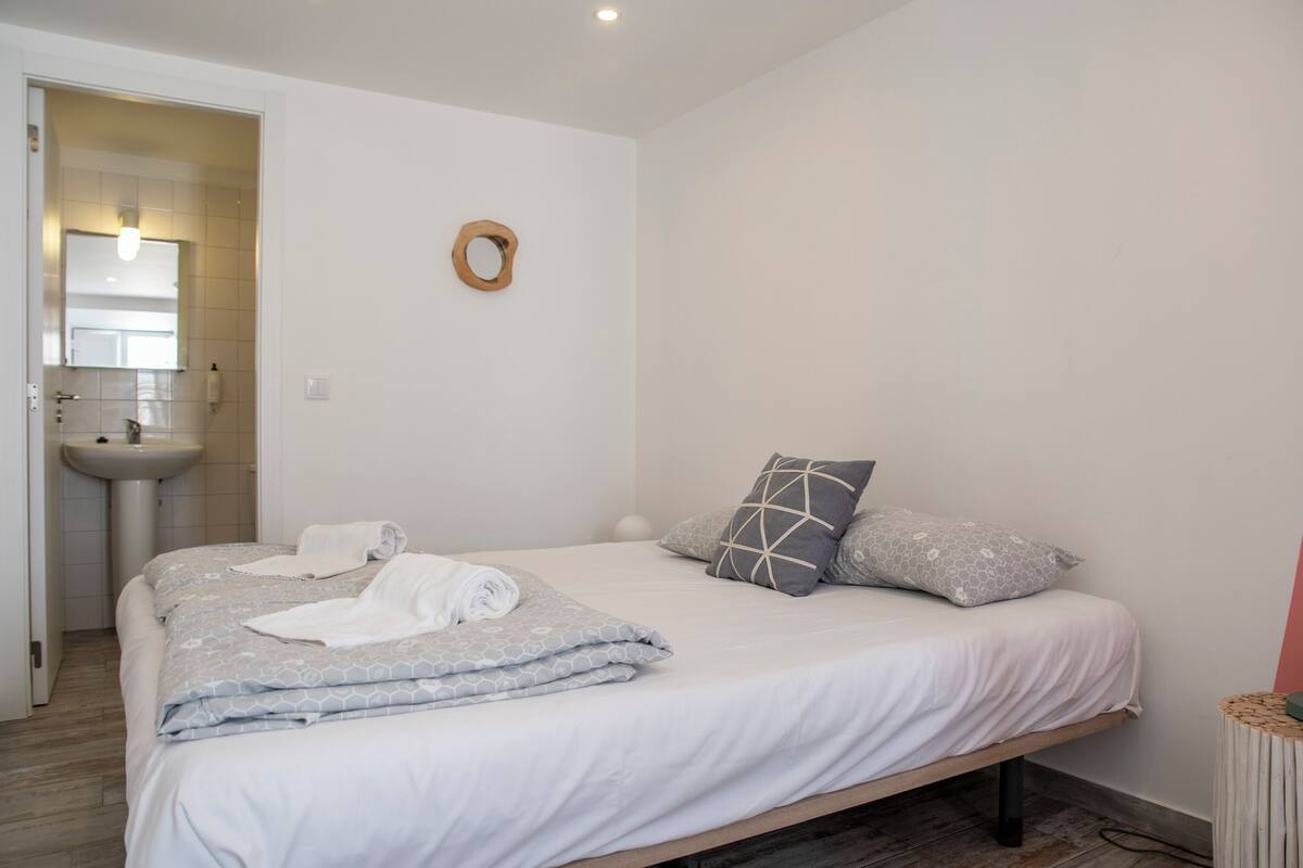 A bedroom with a bed, pillows, a bedside table, and a bathroom with a sink and mirror.