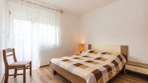 A bedroom with a wooden bed, a chair, a bedside table, and a window with sheer curtains. - Apartments Franjkovic (Novi Vinodolski)