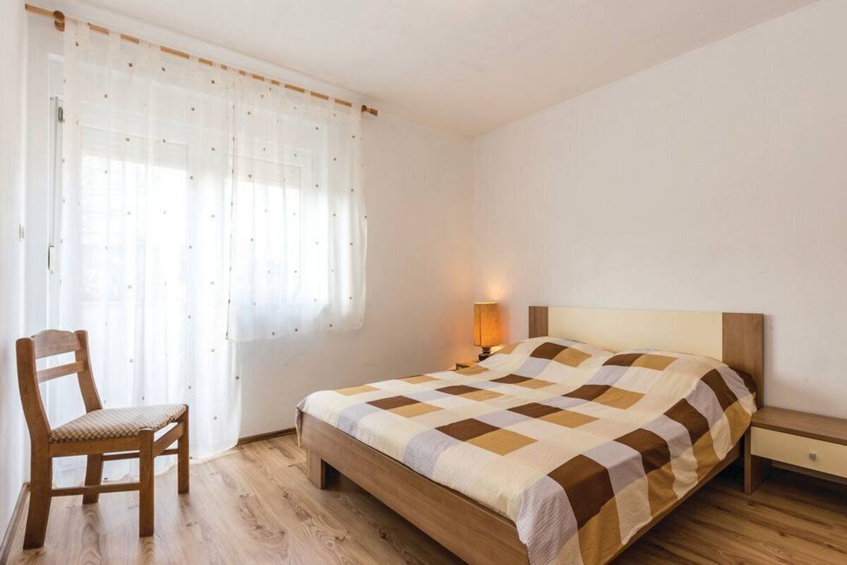 A bedroom with a wooden bed, a chair, a bedside table, and a window with sheer curtains.