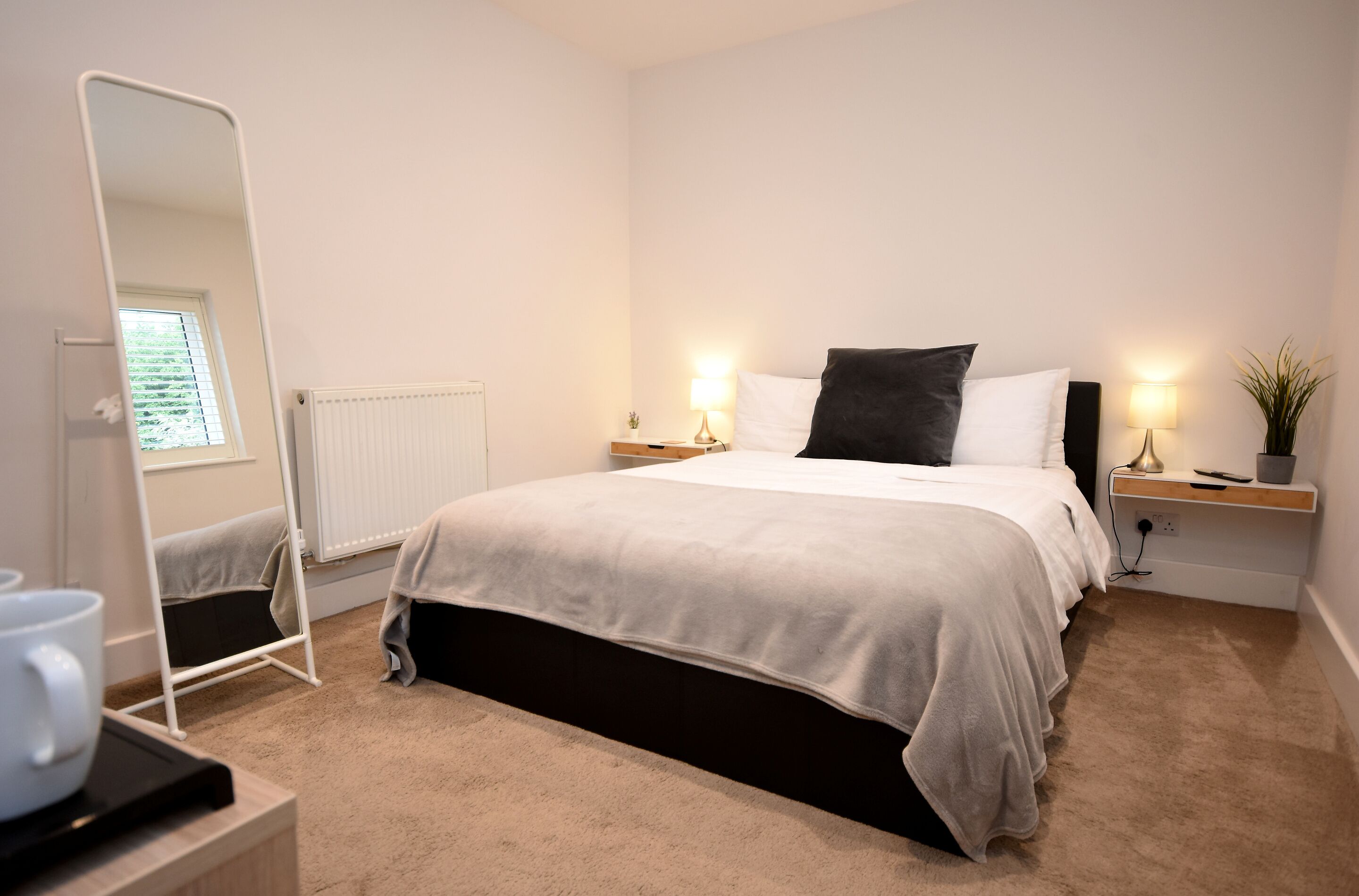 A neatly arranged bedroom with a bed, bedside lamps, a mirror, and a radiator.