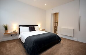 A modern bedroom with a bed, bedside lamps, a nightstand, a wall-mounted radiator, and a door leading to a bathroom. - Apple Tree Place B&B (Tadley)