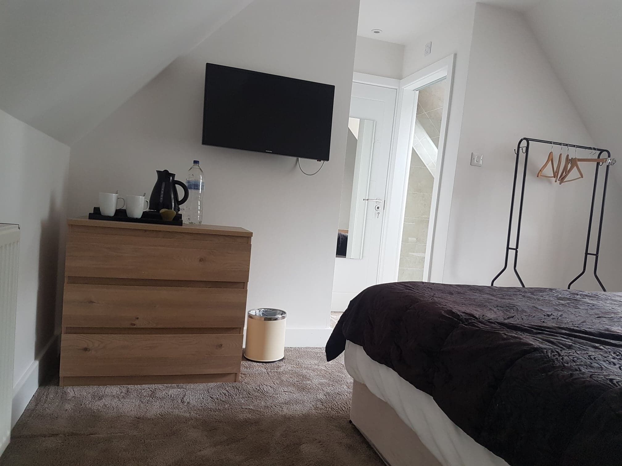 A bedroom with a bed, a wooden dresser, a television mounted on the wall, a radiator, and a door leading to a staircase.