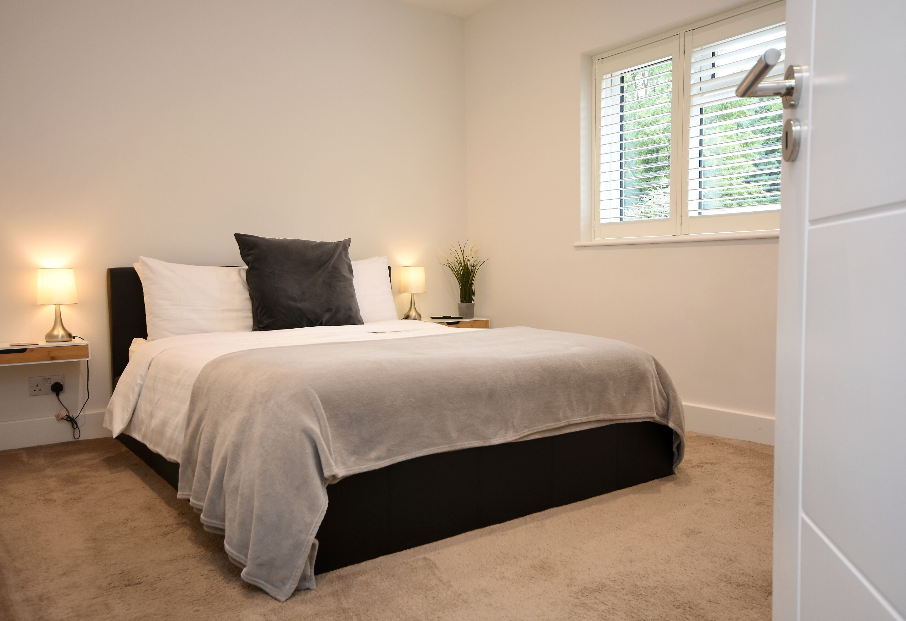 A bedroom with a bed, bedside lamps, a window with blinds, and a door.