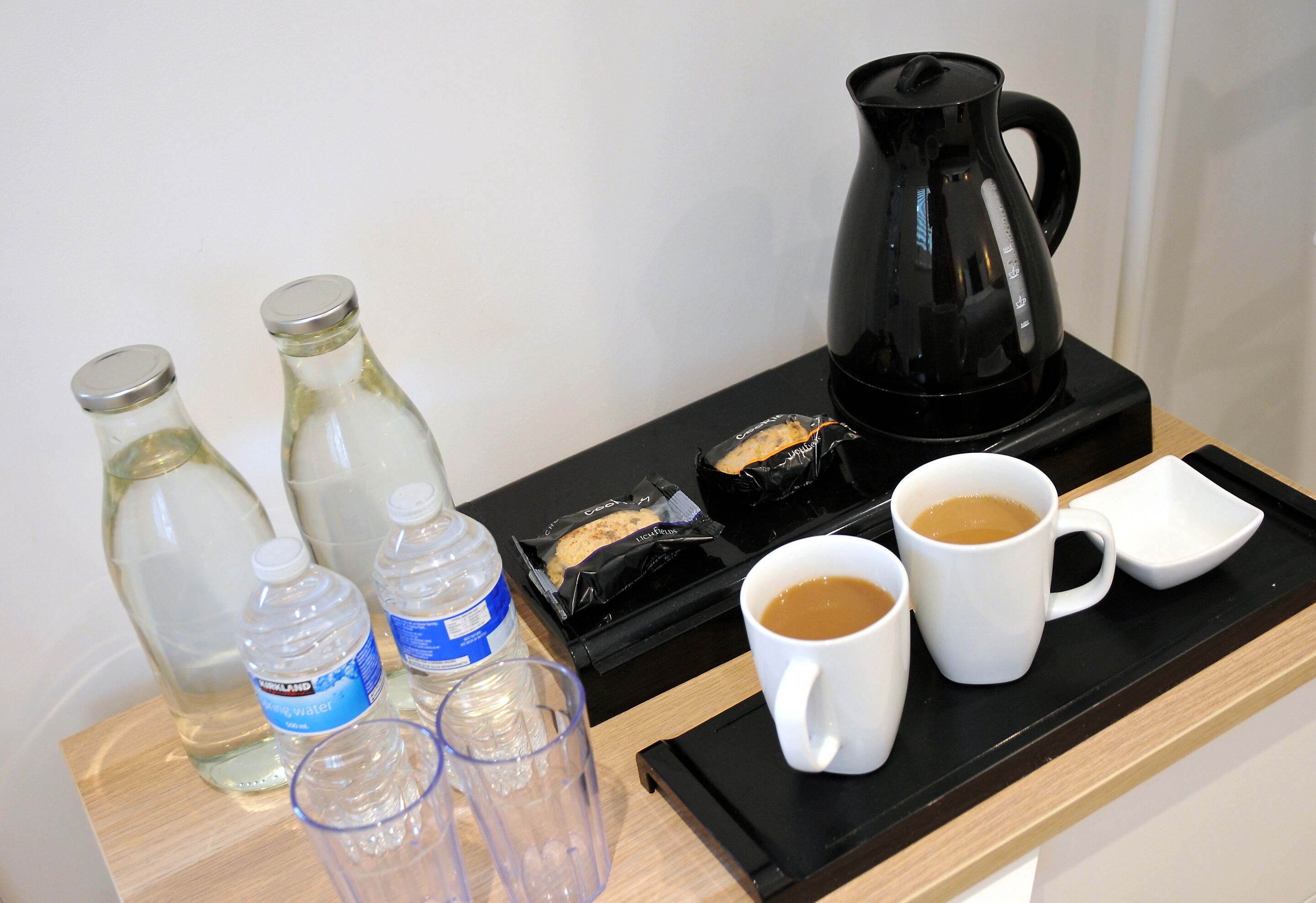 A tray with a black coffee maker, two white mugs, two clear glasses, and two water bottles on a wooden surface.