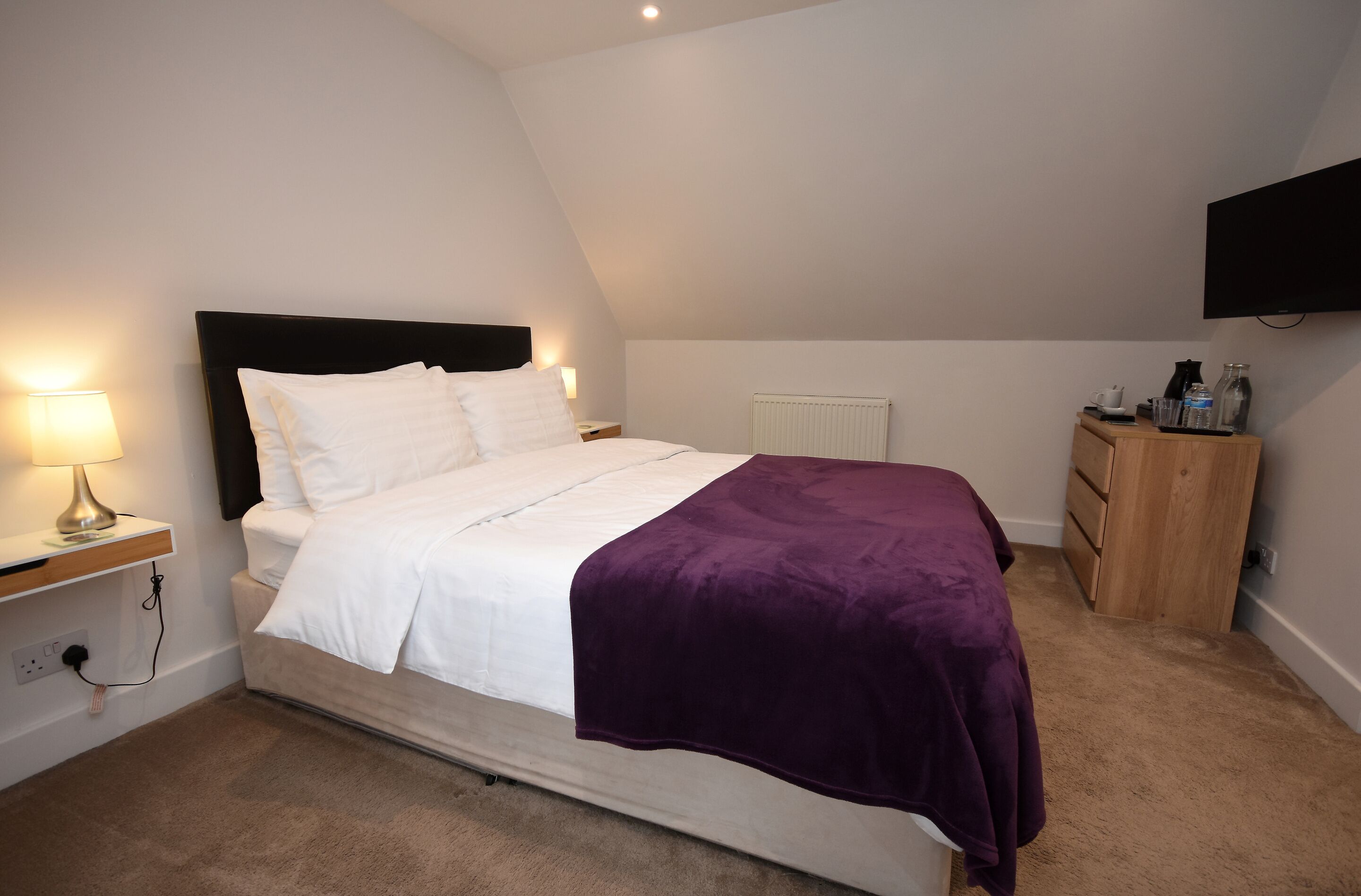 Quarto com cama, mesa de cabeceira com luminária, cômoda e radiador.