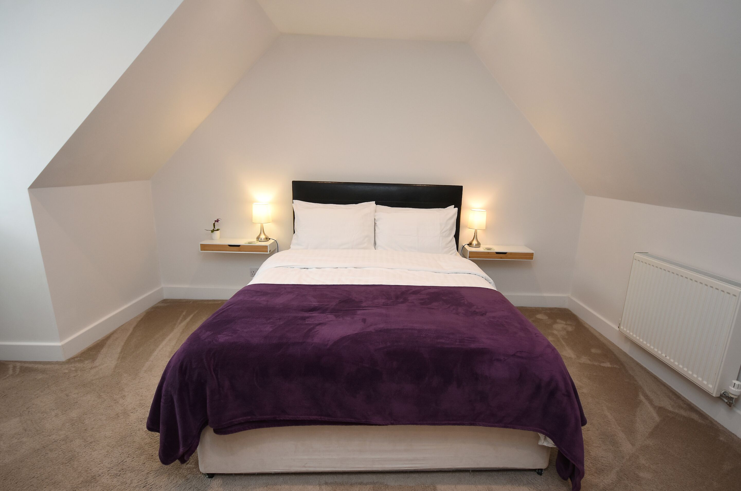 Une chambre à coucher avec un lit, deux tables de chevet avec des lampes, un radiateur et un plafond en pente.
