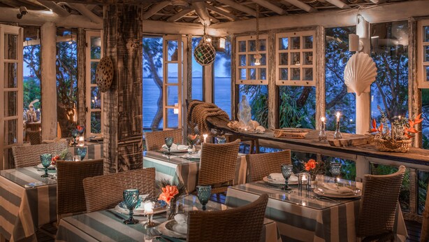 Un restaurante con vista al océano, que cuenta con mobiliario de mimbre, mesas de vidrio y elementos decorativos como conchas y flores.