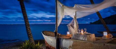A boat with a table set for two, positioned on a sandy beach with palm trees and a calm sea in the background.
