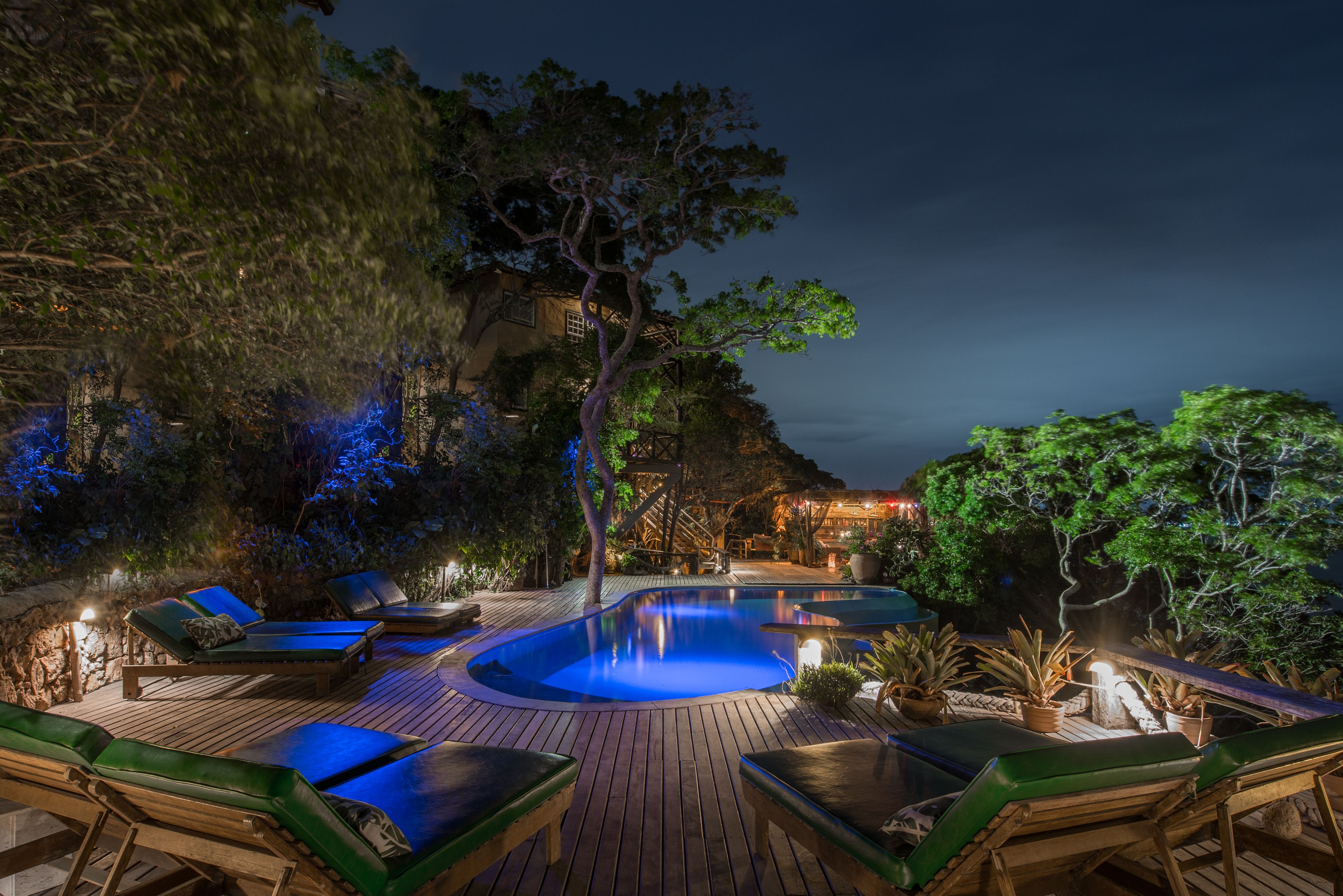 Zona de piscina exterior de noche con sillones de descanso y una terraza de madera.