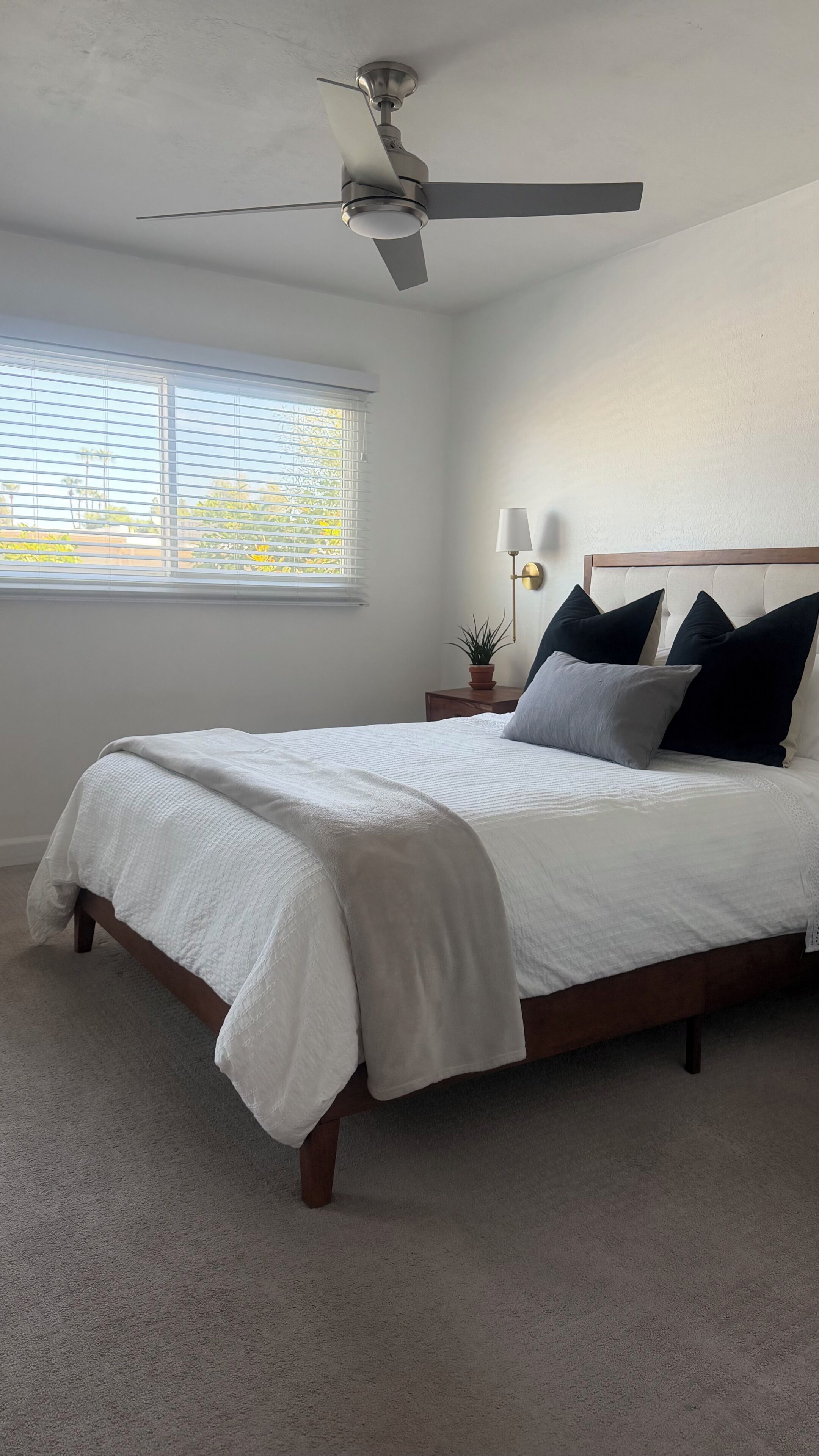 A bedroom with a bed, a ceiling fan, a nightstand with a lamp, and a window with blinds.