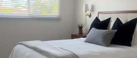 A bedroom with a bed, a ceiling fan, a nightstand with a lamp, and a window with blinds.