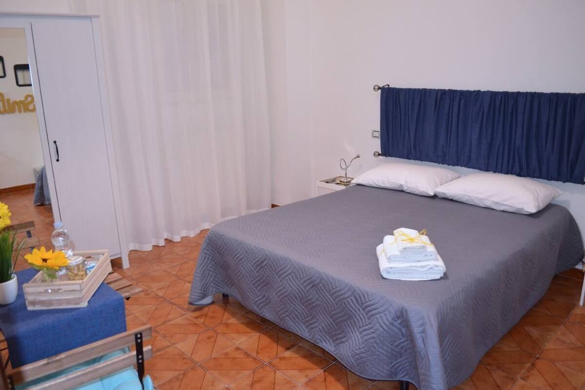 A neatly made bed with a grey bedspread and white pillows, a small bedside table with a lamp, and a window with white curtains.