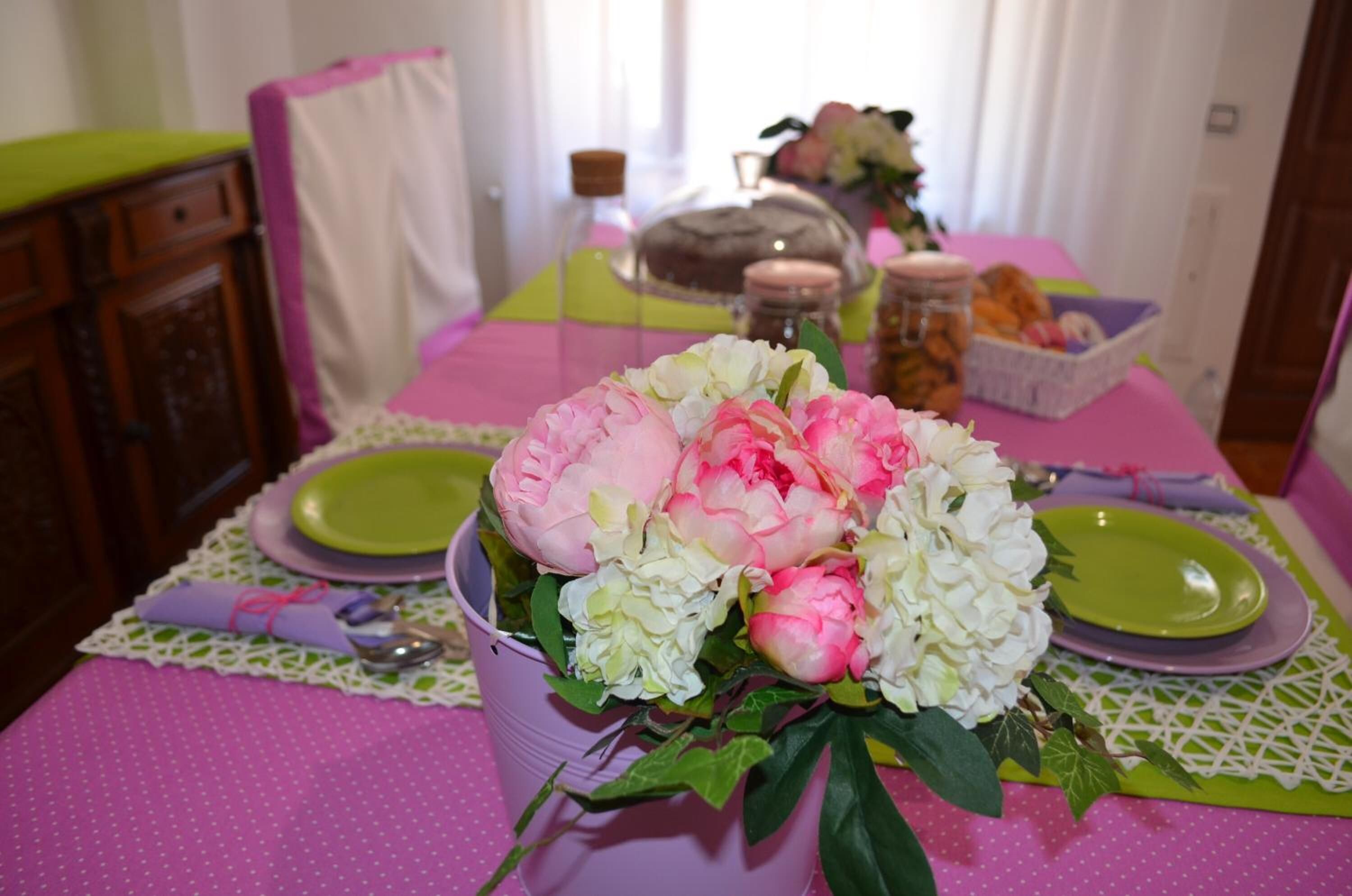 Une table à manger décorée de fleurs roses et blanches, de assiettes vertes et d’un panier rempli de viennoiseries.