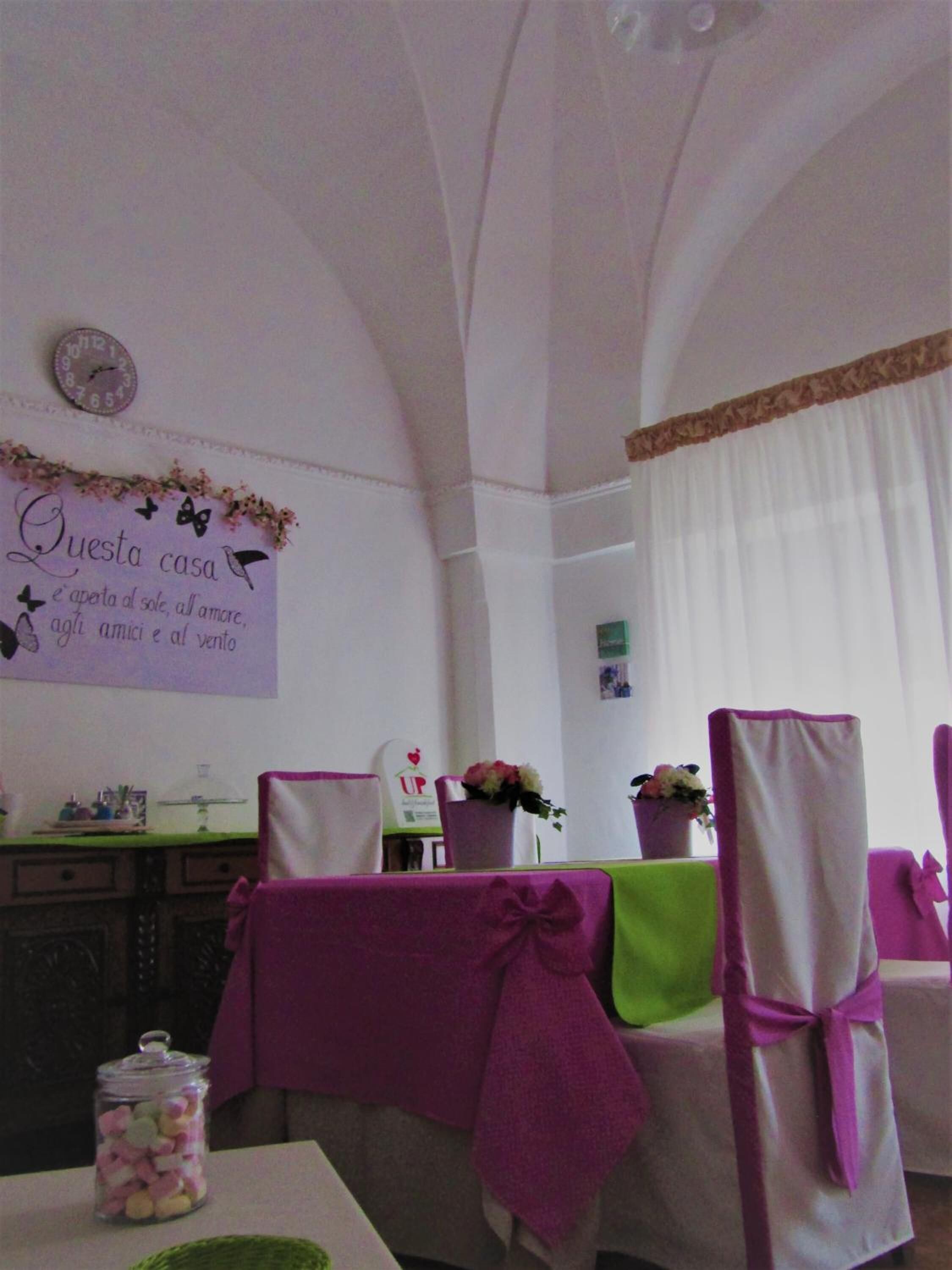 A dining area with a table set for four, chairs with purple sashes, a wall clock, and a sign with Italian text.