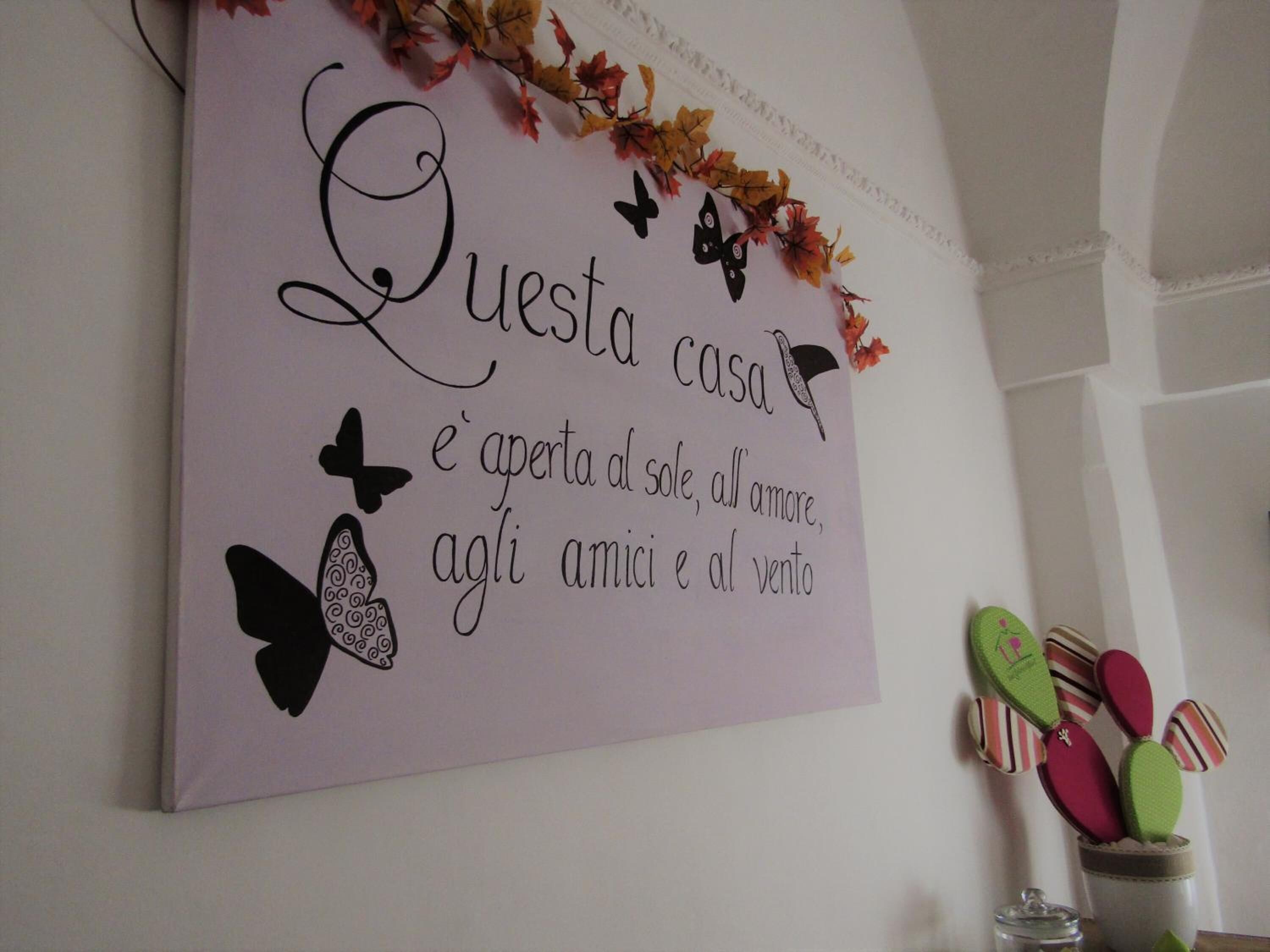 Eine Wand mit einem Schild, auf dem steht: 'Questa casa è aperta al sole, all'amore, agli amici e al vento', verziert mit dekorativen Schmetterlingen und Blättern.