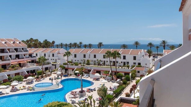 A resort with a central swimming pool, multiple white buildings with red-tiled roofs, and palm trees. - Parque Santiago I 242 - One Bed (Playa de la Américas)
