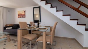 A modern living room with a glass dining table, beige chairs, a sofa, a staircase, and a cityscape painting on the wall. - Parque Santiago I 242 - One Bed (Playa de la Américas)
