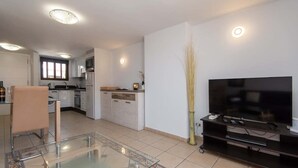 A modern living room with a glass coffee table, a flat-screen TV on a black stand, and a kitchen in the background. - Parque Santiago I 242 - One Bed (Playa de la Américas)