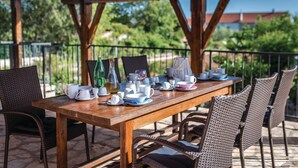 An outdoor dining area with a wooden table set for tea service, wicker chairs, and a view of greenery and buildings in the background. - Amazing home in Cista Velika (Cista Velika)
