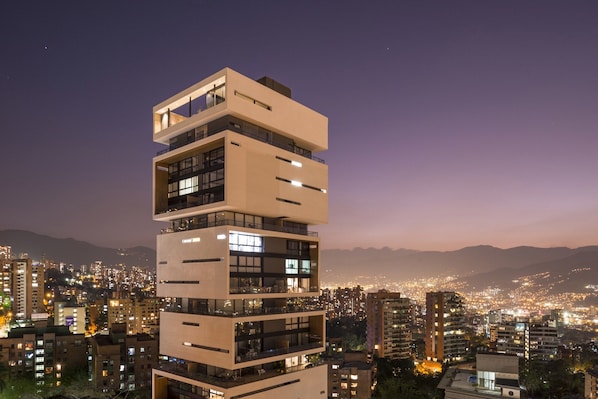 Um edifício com vários andares, varandas e janelas amplas, com o horizonte da cidade ao fundo, à noite.