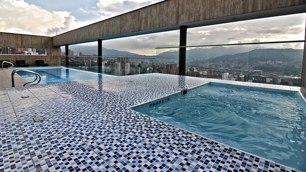 Une piscine à débordement située sur le toit, offrant une vue sur la ville, avec une balustrade en verre et un sol carrelé.
