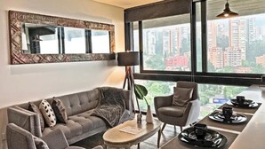 Un salon moderne avec un canapé gris, une petite table et un fauteuil. Une grande fenêtre offre une vue sur de hauts immeubles.