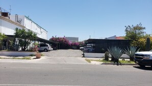 Vista de la calle con coches aparcados, un edificio de fachada blanca y un cielo azul despejado.